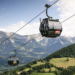 Zwei Gondelbahnen am Stahlseil mit grünen Wiesen und einer imposanten Bergkette im Hintergrund