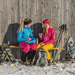 Zwei Frauen mit Schneeschuhwander-Ausrüstung auf Bank an Holzwand gelehnt