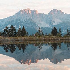 Radfahrer auf Wiesenweg vor See in dem sich Berge spiegeln